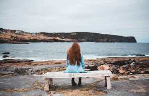 woman with red hair in teal jacket on bench by water