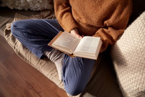 Woman in a sweater and jeans sitting cross legged on couch with an open book