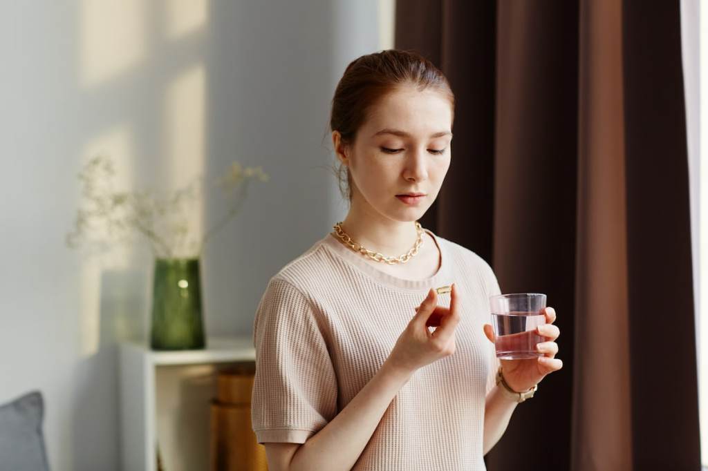 Woman in a tan shirt holding a small pill and a glass of water in her hands