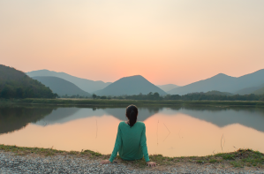 A woman sits alone by a still lake at sunset, looking out toward a mountain landscape.