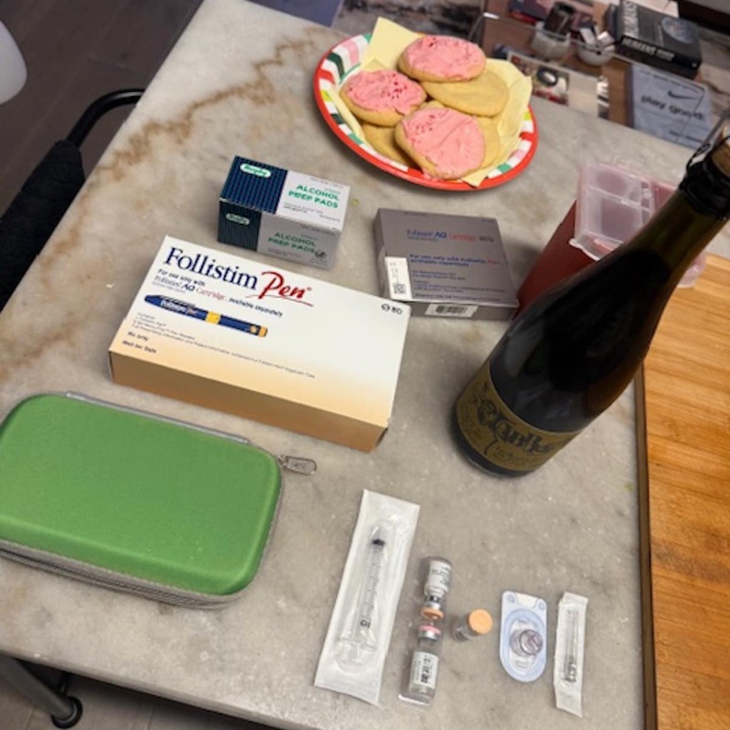 A marble countertop with pink sugar cookies, a Folllistim Pen, and a bottle of champagne as a woman prepare to inject hormones in preparation of freezing her eggs