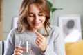 Woman in a gray sweater holding a glass of water taking a white pill