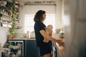A tired mother sighs in the kitchen while holding her crying newborn and preparing breakfast, representing burnout