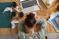 Overwhelmed young woman with her head down on a messy office desk with laptop, coffee cups, and crumpled papers, top view